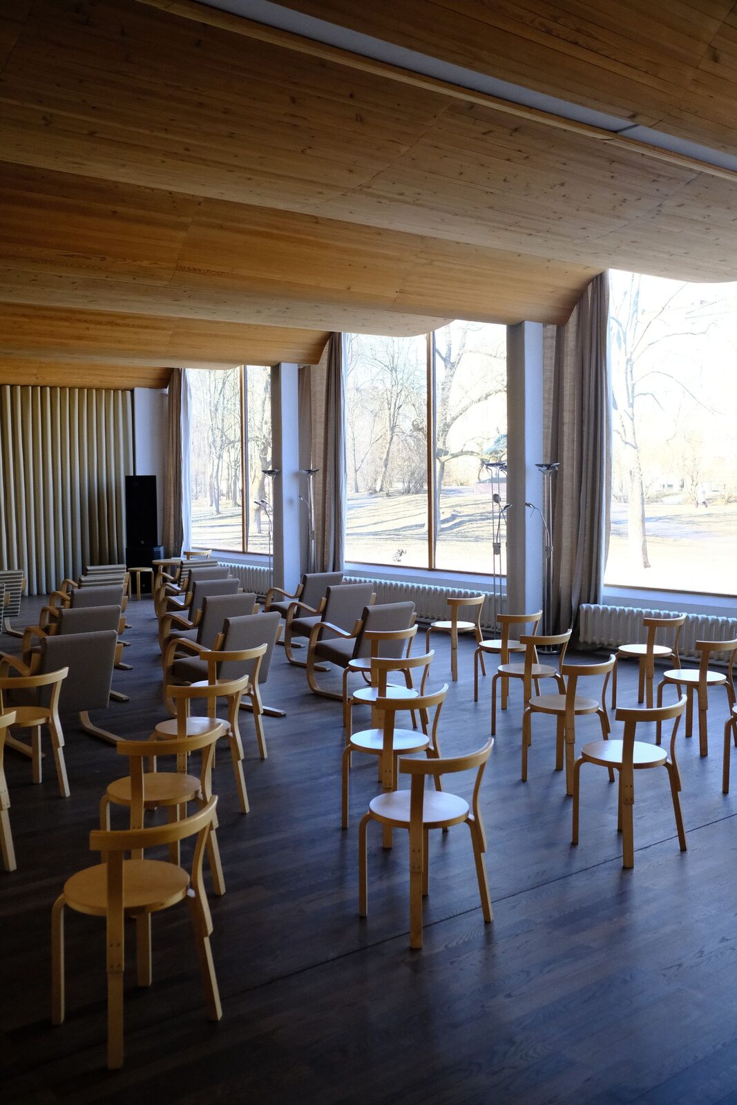 La grande salle vide, plafond de bois et verrière ouverte sur le parc, chaises en attente